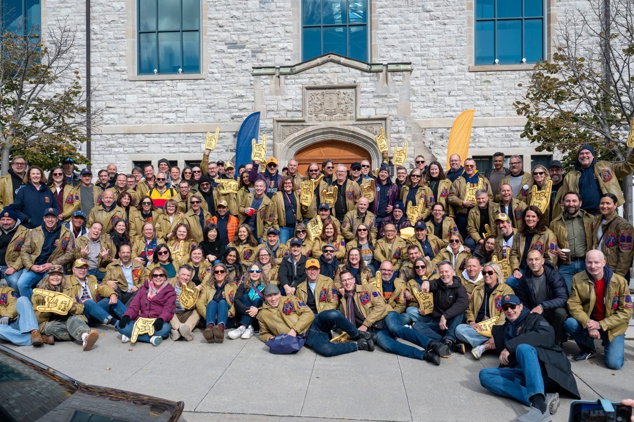 Sci'96 classmates in gold jackets with foam 'Sci 96 Rocks Hoco' fingers, at Mitchell Hall during Homecoming 2026